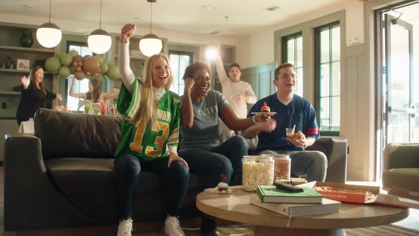 A diverse group of friends in team jerseys erupts in celebration while watching a sports game at a lively house party