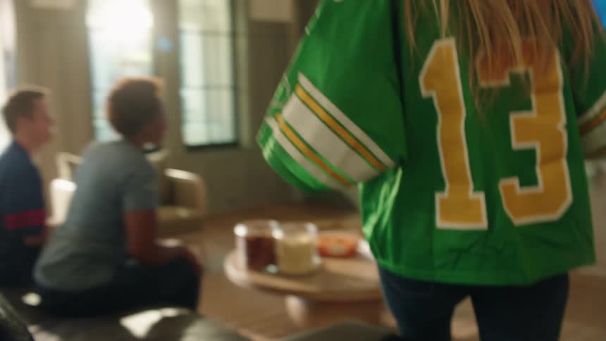 A woman in a football jersey happily brings cupcakes to her friends while they watch a game on TV together
