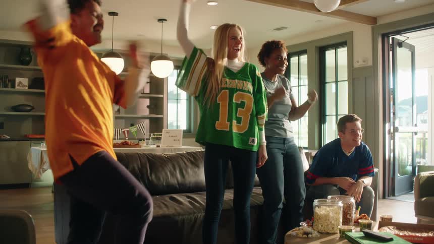 Excited sports fans in team jerseys leap up and celebrate a victory at a watch party while their dejected friend mourns his team's loss
