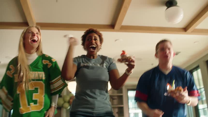 Three diverse friends wearing football jerseys enthusiastically celebrate a victory while watching a game at home, jumping and cheering in slow motion