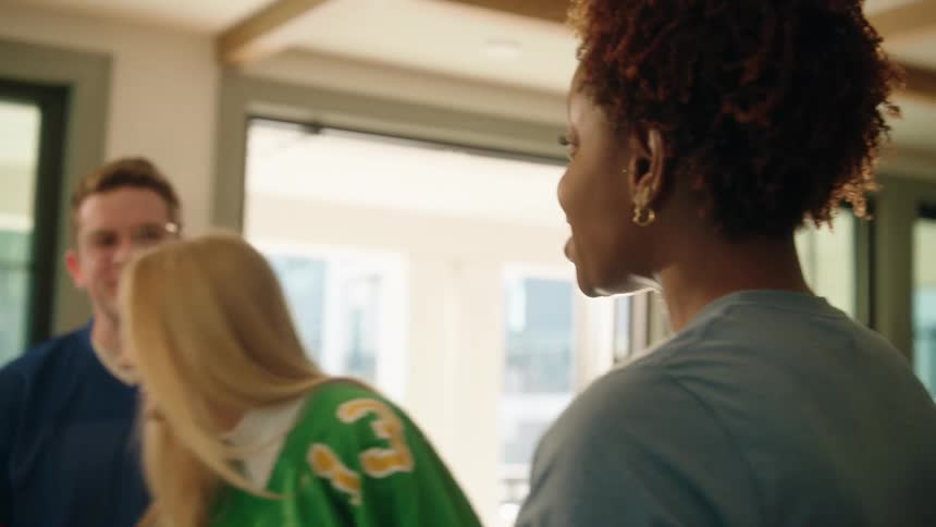 A group of diverse friends in football jerseys laugh and grab snacks from a food table at a game day party