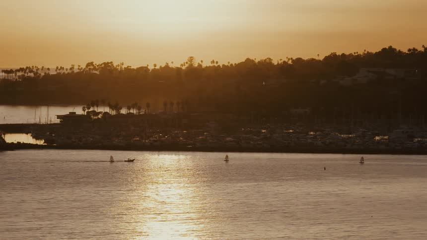 A serene wide shot of a coastal harbor at sunset, with small boats sailing on the golden, shimmering water
