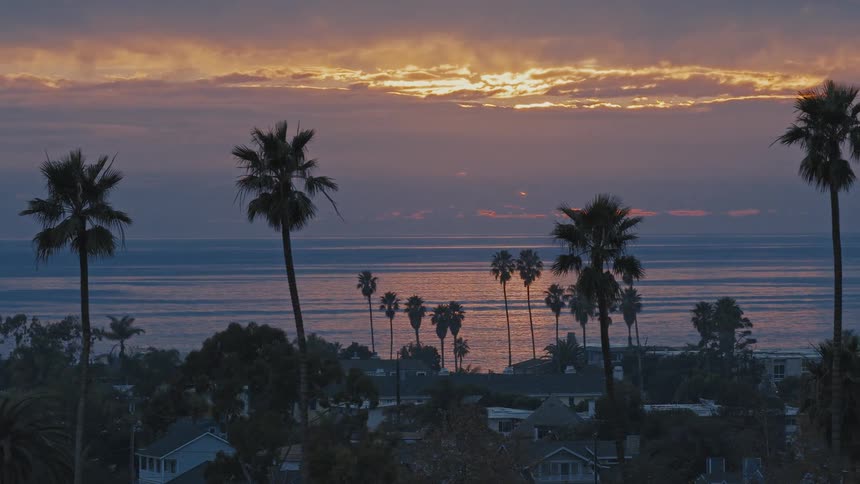 A colorful sunset over the ocean illuminates the sky behind silhouetted palm trees and a quiet beach town