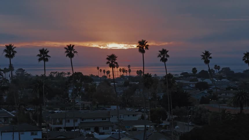 A beautiful golden sunset reflects on the ocean behind a California beach town with silhouetted palm trees