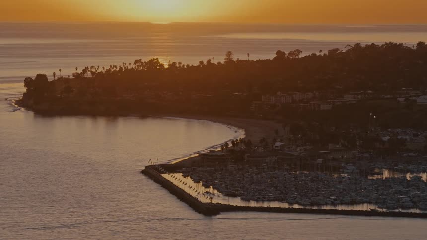 A serene high-angle view shows the golden sunset over the ocean, illuminating a bustling harbor and the picturesque coastline