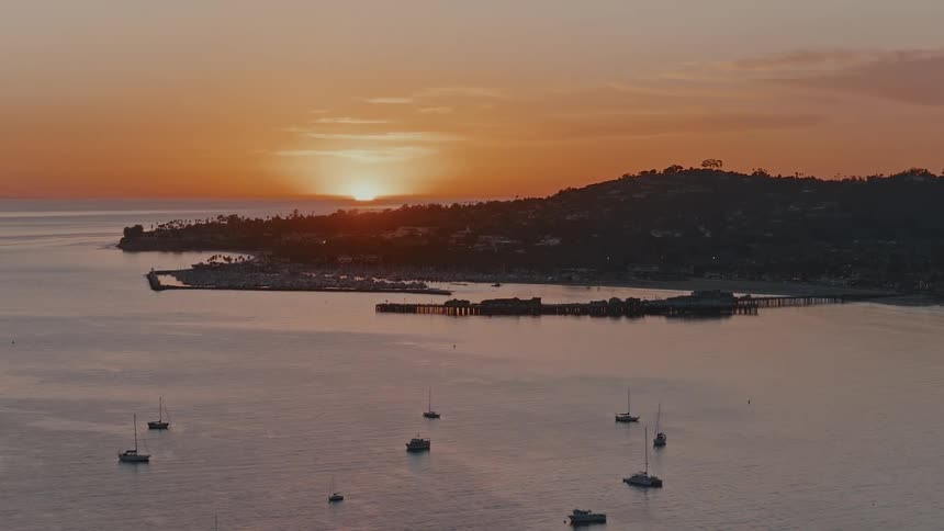 A serene sunset casts an orange glow over a tranquil bay where sailboats are anchored near a coastal town and pier