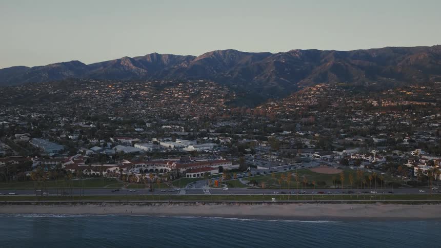 A stunning aerial view captures the scenic coastline of Santa Barbara, California, slowly moving towards the city with the Santa Ynez Mountains in the background at sunset