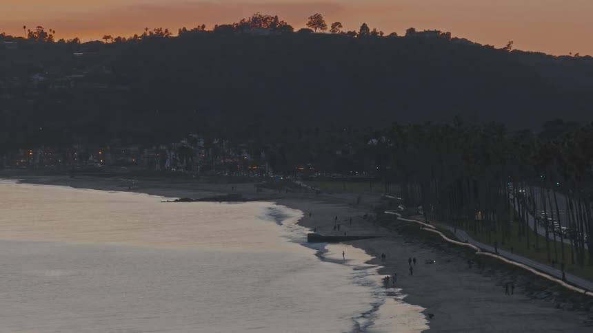 An elevated view captures the tranquil Santa Barbara coastline at dusk, with people walking on the beach and a palm tree-lined road