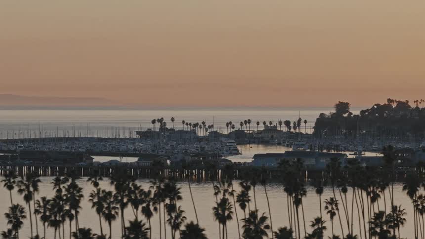 An idyllic evening view of the Santa Barbara marina with boats, a pier, and silhouetted palm trees under a warm sunset sky