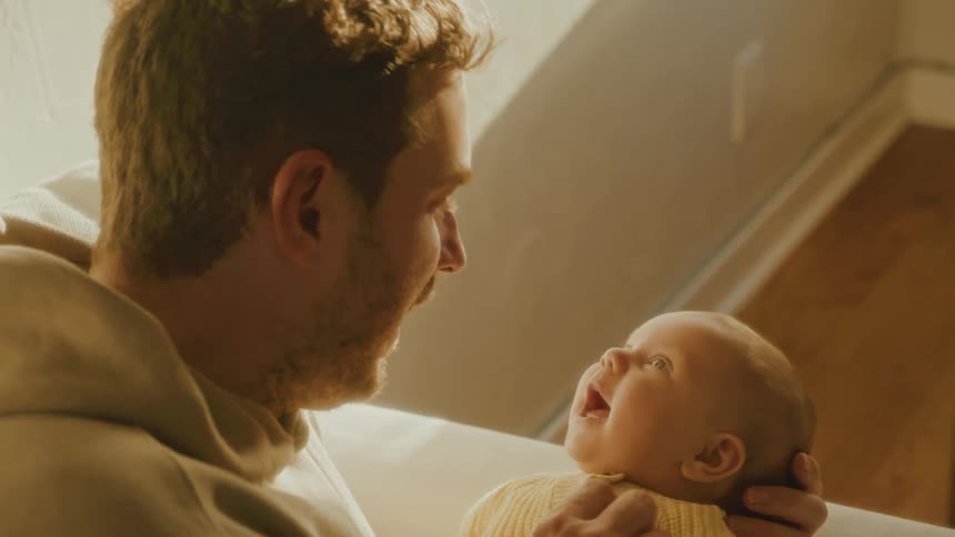 A new father lovingly holds his newborn baby who smiles with joy in a warm, sunlit room