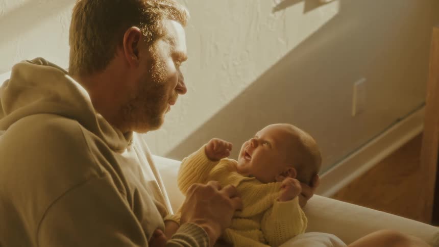 A loving father holds his smiling newborn baby on his lap while sitting on a couch in warm sunlight