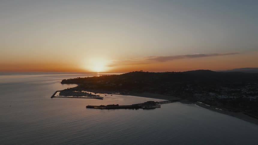 A wide aerial shot captures a coastal city with a pier and harbor silhouetted against a beautiful golden sunset over the calm ocean
