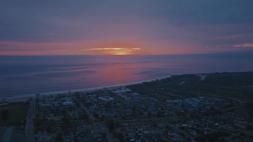 A stunning aerial view captures a vibrant pink and purple sunset reflecting on the calm ocean by a coastal town