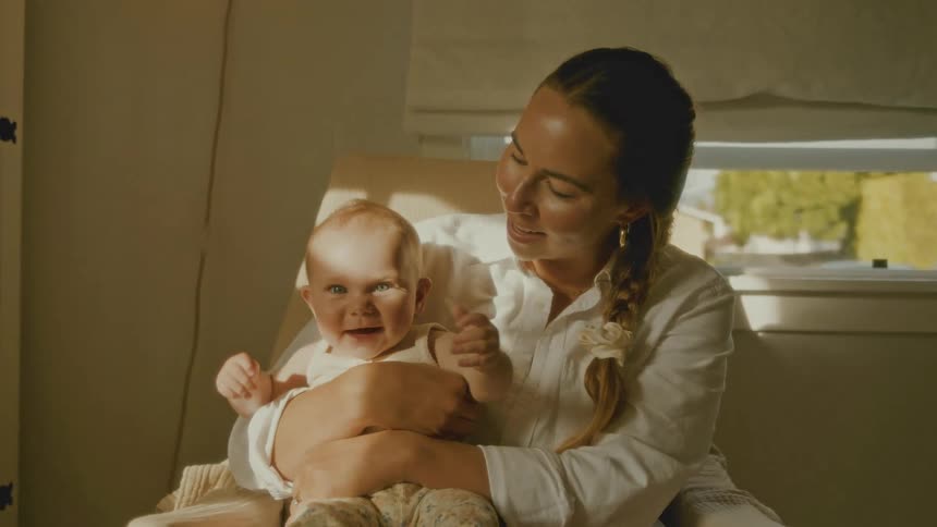 A loving mother holds her happy baby in her arms while sitting in a chair bathed in warm sunlight from a nearby window