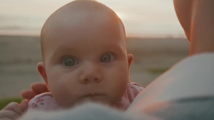 An adorable baby with wide, curious eyes looks over its mother's shoulder during a beautiful sunset on the beach