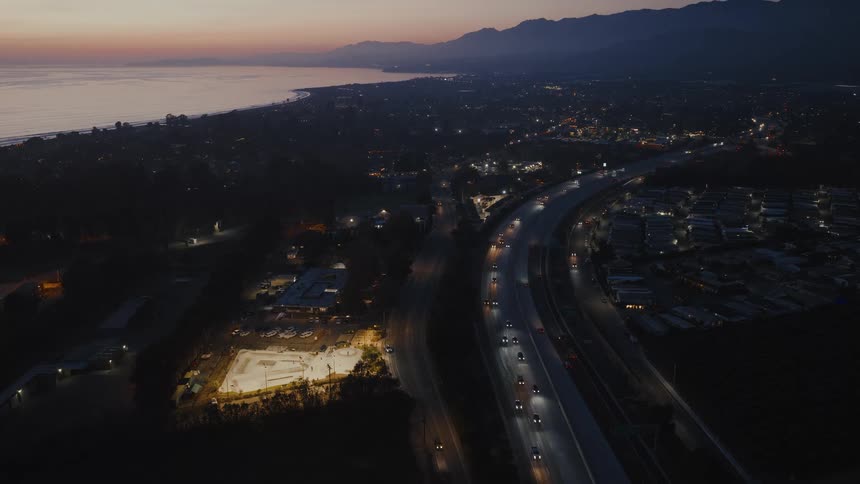 Aerial view of a coastal city at dusk with traffic flowing on the highway next to a brightly lit skatepark and the ocean