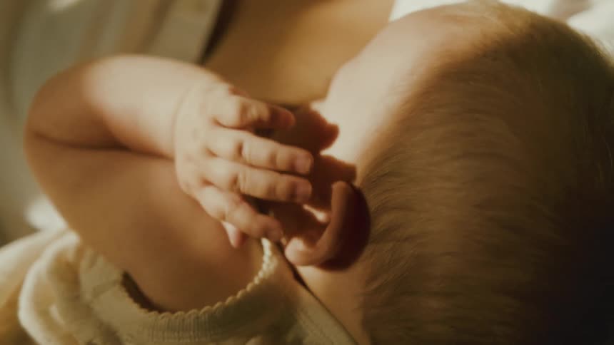 A tender close-up from behind a newborn baby as they breastfeed in their mother's arms in a softly lit room