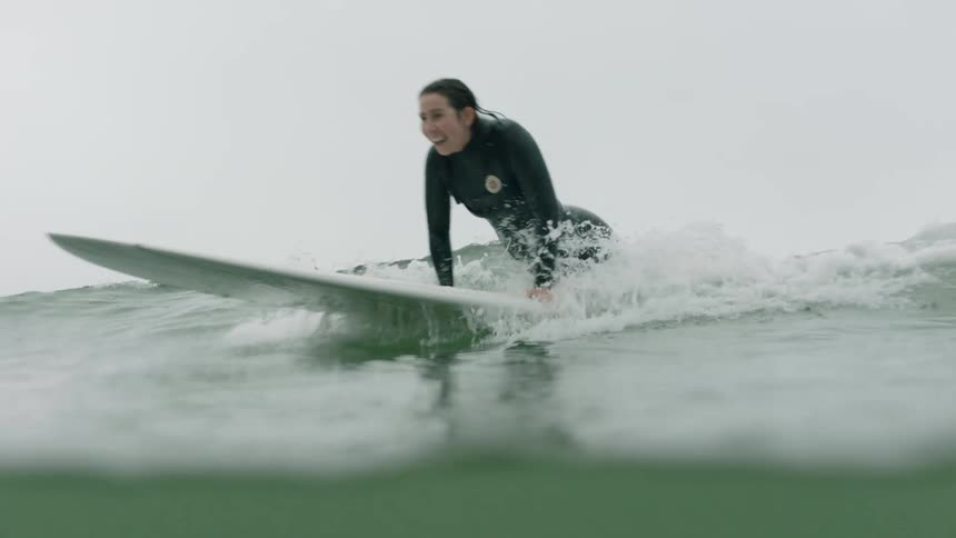 A smiling woman in a wetsuit catches an ocean wave on her surfboard in slow motion
