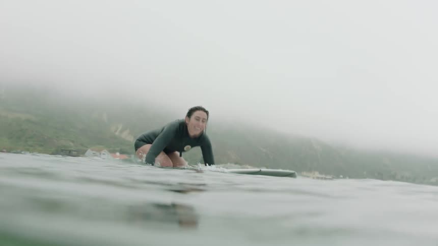 Slow motion water-level view of a smiling female surfer in a wetsuit riding gentle waves on a summer coastal morning