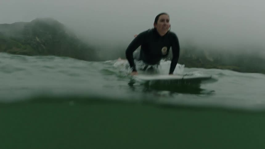 In a slow-motion split-level view, a female surfer in a wetsuit paddles on her board through the foggy ocean