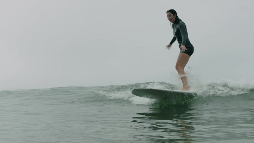 A female surfer gracefully pops up and rides a gentle wave in slow motion on a summer day, with the camera dipping underwater as she passes