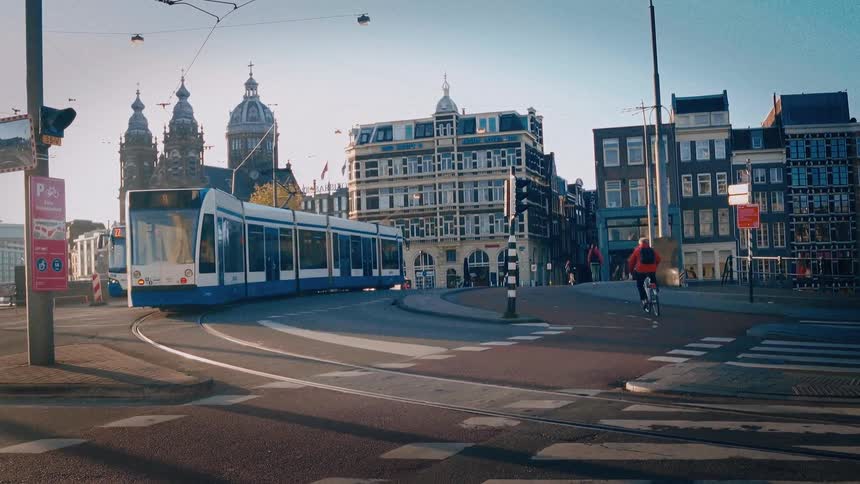 A man rides his bicycle through a quiet intersection in Amsterdam as a tram passes, revealing the city's historic architecture in the morning light