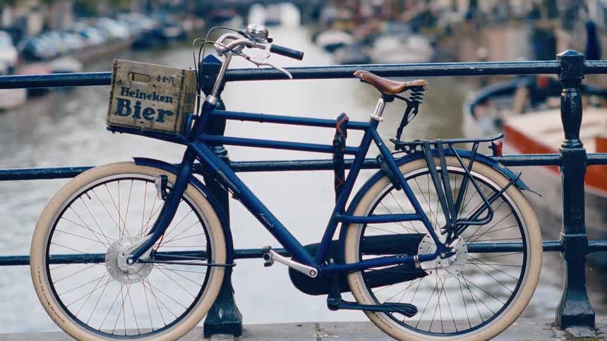 A classic blue bicycle with a Heineken beer crate rests on a bridge overlooking a scenic Amsterdam canal