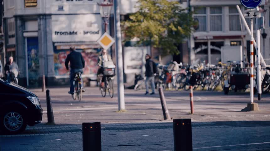 A slow-motion, out-of-focus shot shows cyclists, pedestrians, and cars moving through a sunlit city intersection