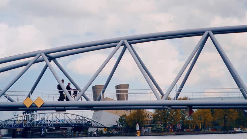 Side-view tracking shot of pedestrians and a cyclist crossing a modern grey truss bridge over a city river on a cloudy day