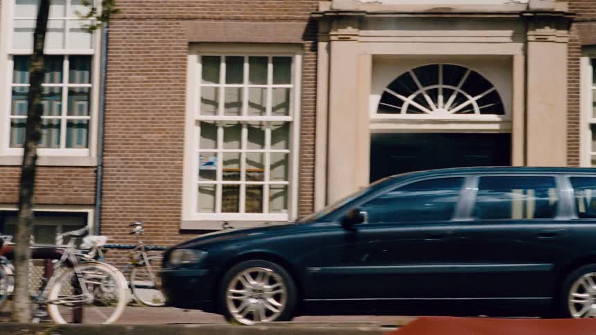 Parallax view of a classic Amsterdam street featuring brick townhouses, parked bicycles, and people passing by on a sunny day