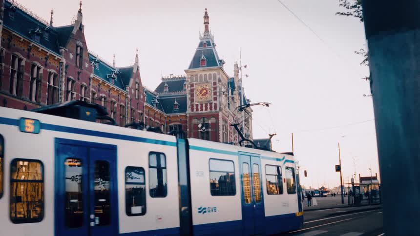 A modern city tram slowly moves past the historic Amsterdam Centraal Station at dusk