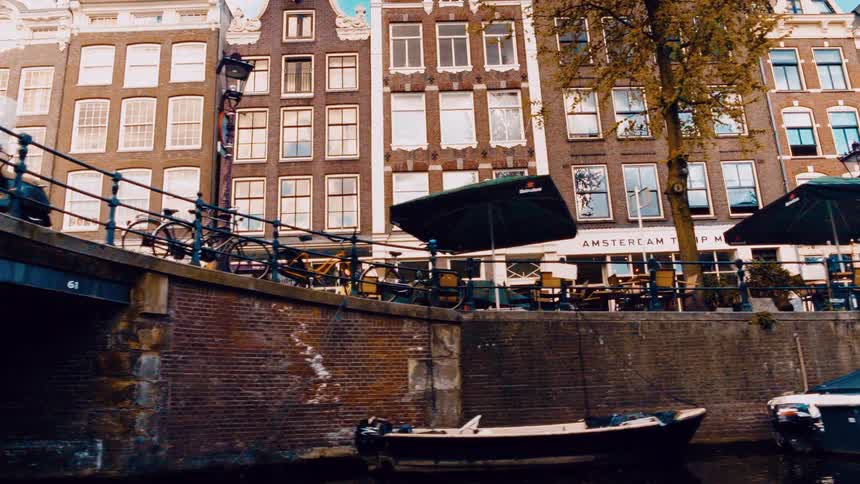 A boat's point of view gliding along an Amsterdam canal, passing classic Dutch houses, a waterside cafe, and a brick bridge