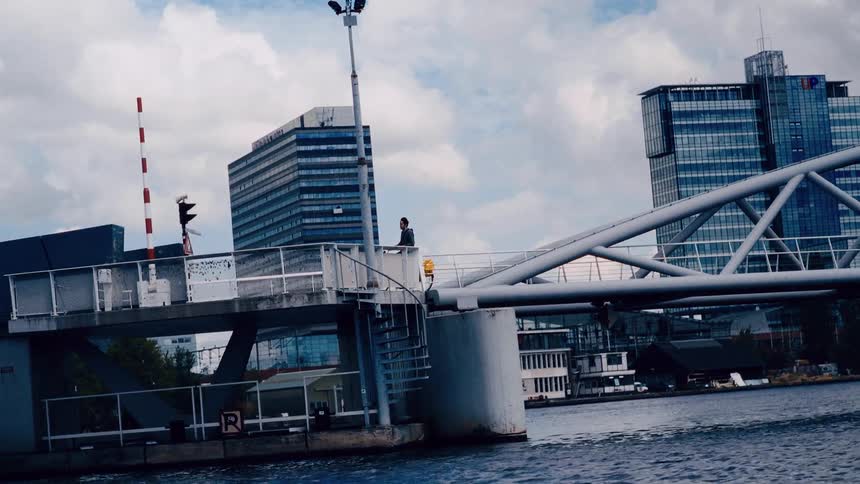 A cyclist pedals across a modern drawbridge in slow motion with a cityscape in the background