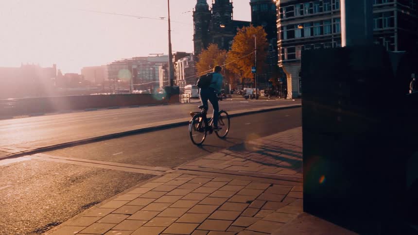 A man with a backpack rides his bicycle away from the camera down a city street during a cinematic golden hour sunset