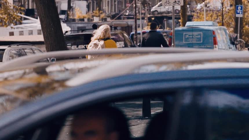 A woman in a vibrant yellow jacket rides her bicycle down a bustling city street in Amsterdam in slow motion