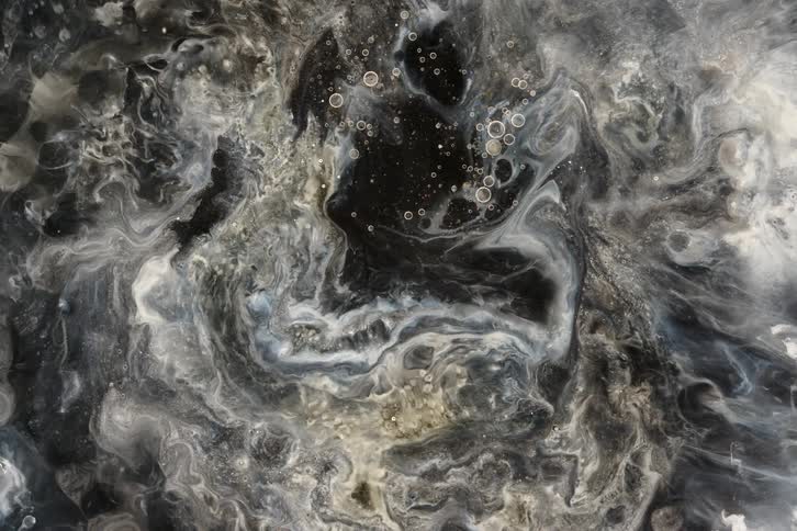 An ethereal top-down shot of abstract black, white, and grey liquids swirling slowly, resembling a distant galaxy or a chemical reaction