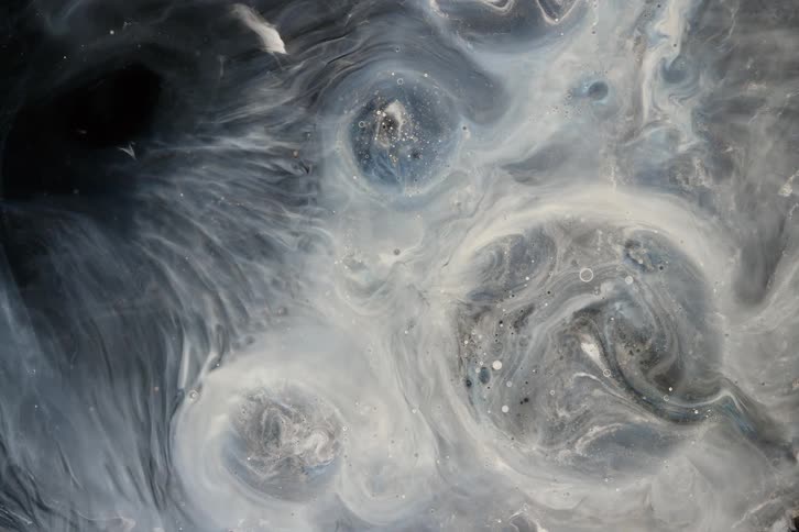 An abstract top-down view of white and gray liquids with glitter swirling slowly to create a cosmic, nebula-like pattern