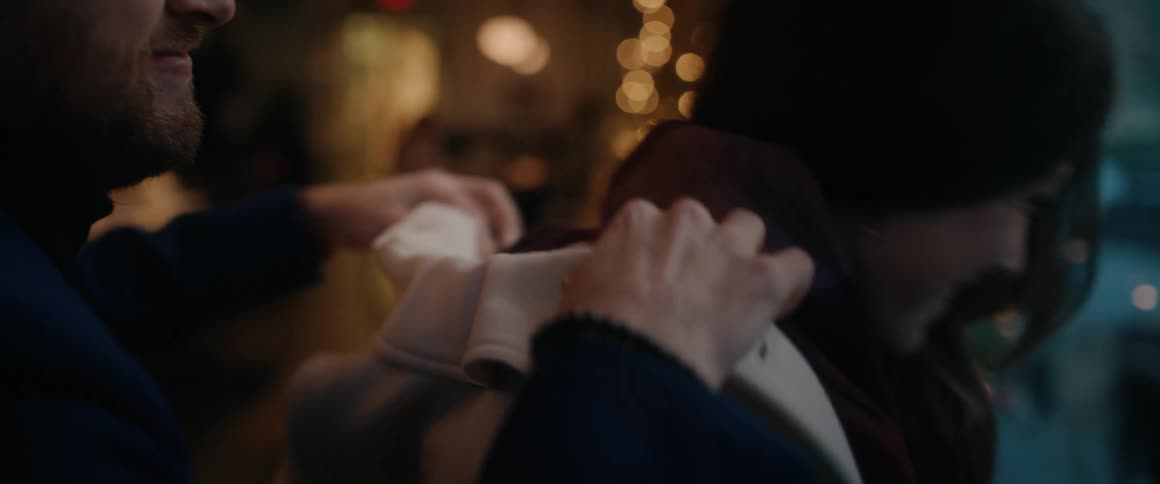 Close up of a man lovingly placing a coat on a woman's shoulders in a warm, festive environment