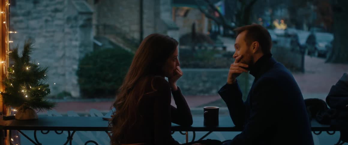 A man and a woman sit at a cafe table by the window, engrossed in a romantic conversation at dusk