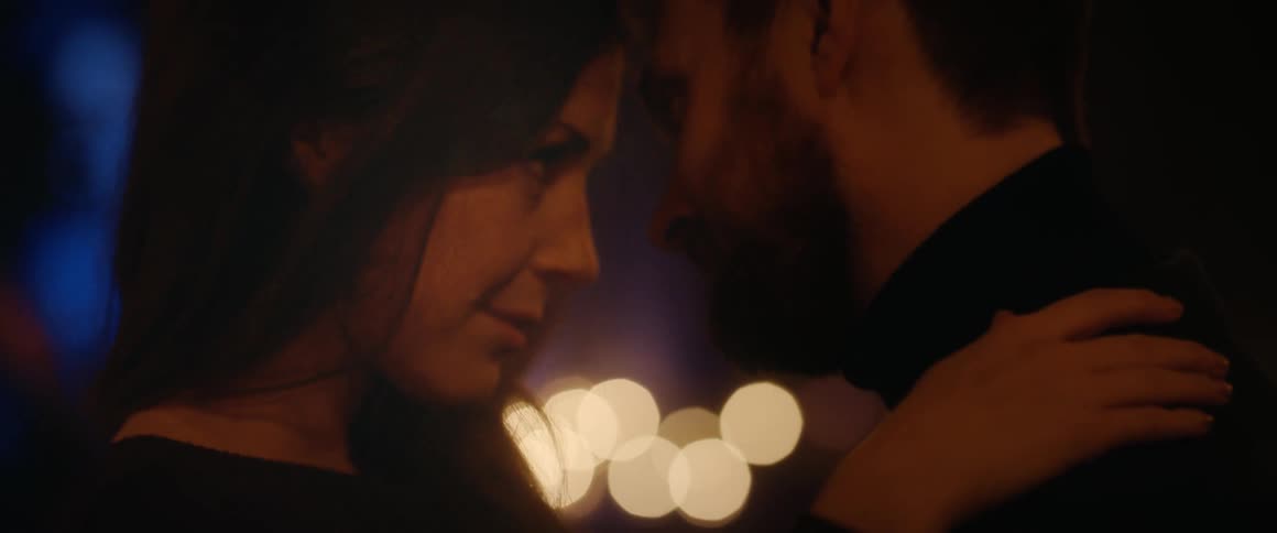 A romantic couple holds each other close, their foreheads touching in an intimate moment with soft bokeh lights in the background