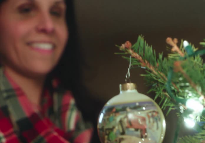 A smiling woman carefully hangs a vintage-style ornament on a Christmas tree, with the focus shifting to the bauble