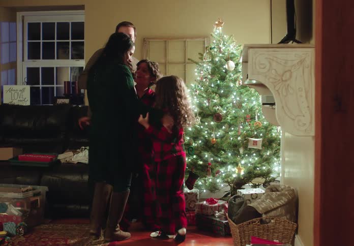 A mother returns home for the holidays and is greeted with a surprise hug from her family by the Christmas tree
