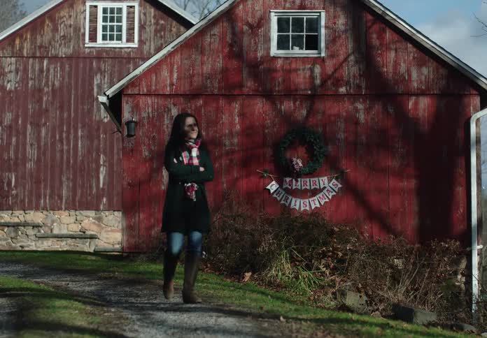 A woman in a cozy winter coat and scarf walks in slow motion past a rustic red barn decorated for the holidays