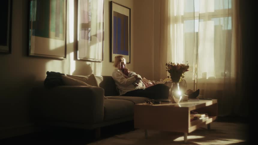 A grandmother lovingly holds her newborn grandchild, resting together on a sofa bathed in the warm light of the afternoon sun