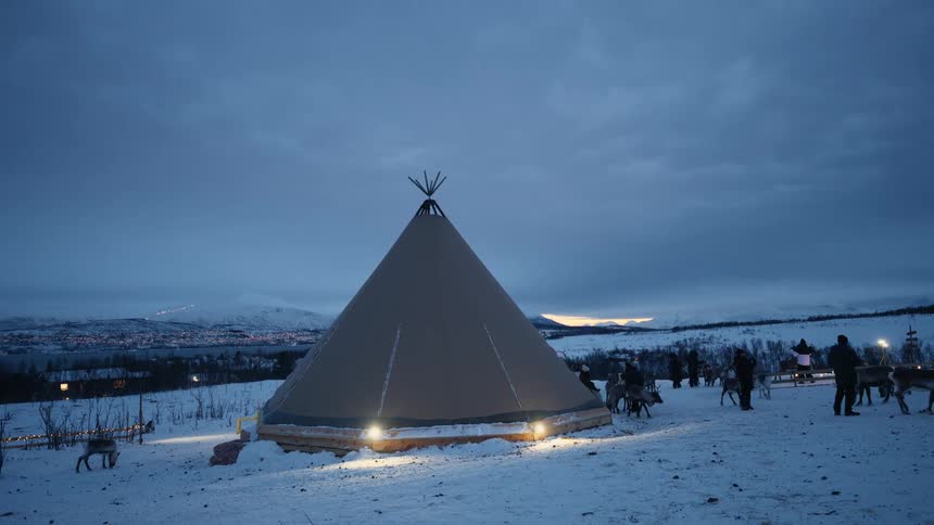 A traditional Sami lavvu tent is illuminated at dusk in a snowy landscape as people and reindeer gather nearby, with a glowing town visible in the distance