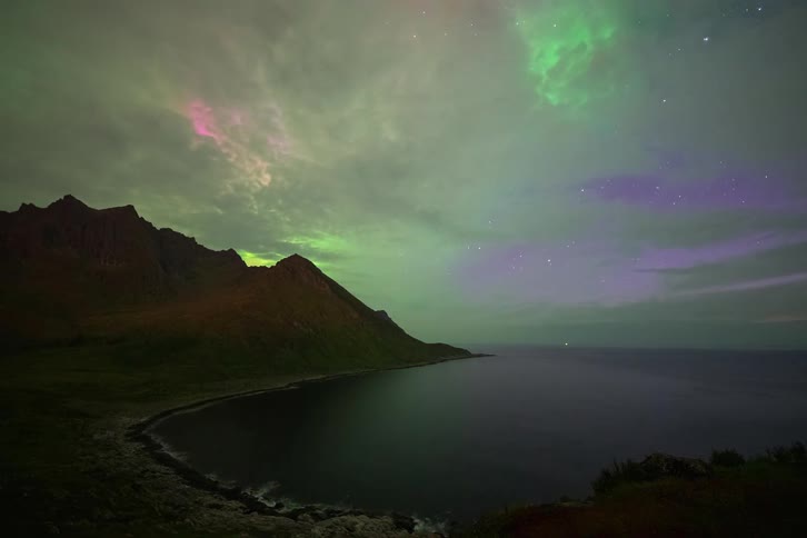 A stunning time-lapse shows the vibrant green and purple Aurora Borealis dancing in the cloudy night sky over a remote mountainous coastline