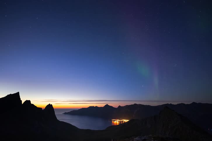A stunning timelapse shows the green and purple Aurora Borealis dancing in the starry night sky over a mountainous fjord and an illuminated coastal town
