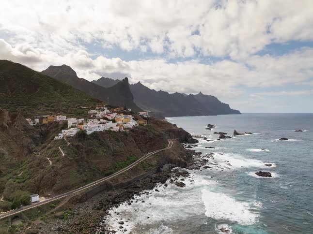 An aerial view pulls back to reveal a stunning coastline with a winding road where cars drive past a small village perched on the rugged cliffs