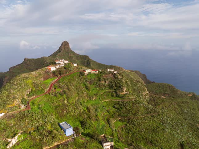 A stunning aerial pullback shot reveals a secluded village on a verdant mountainside above the ocean under a cloudy sky
