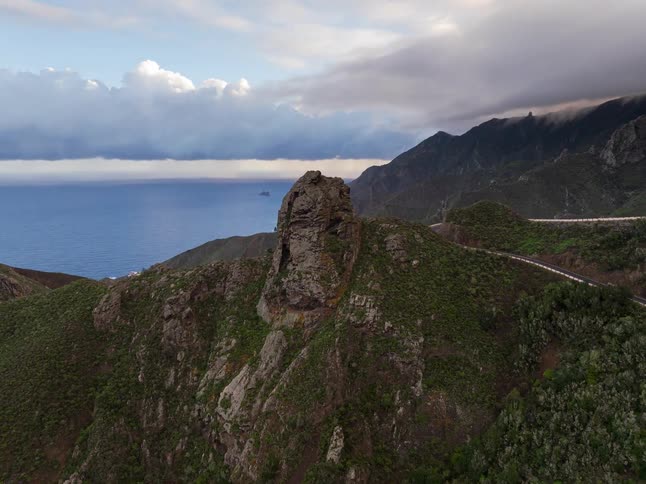Aerial footage reveals a dramatic green mountain landscape with a winding road overlooking the ocean under a cloudy sky